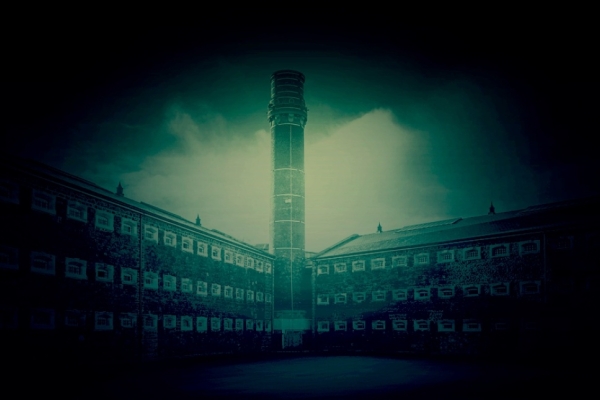 A green-blue tinted image of a tall, external brick tower overlooking three-storey Y-shaped brick prison wings containing dozens of small windows